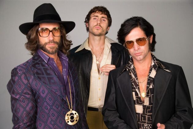 Na imagem, três homens com expressões sérias aparecem vestidos com camisas chamativas e blaseres antigos da década de 70 com correntes de ouro e óculos estilo avidor.