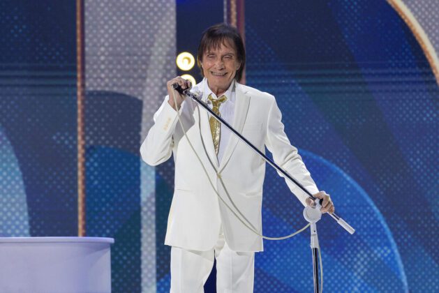 Roberto Carlos aparece sorrindo de terno branco com gravata dourada, segurando o microfone.