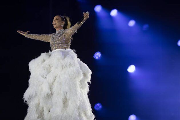 Ivete Sangalo está de braços abertos e vestido branco e prateado