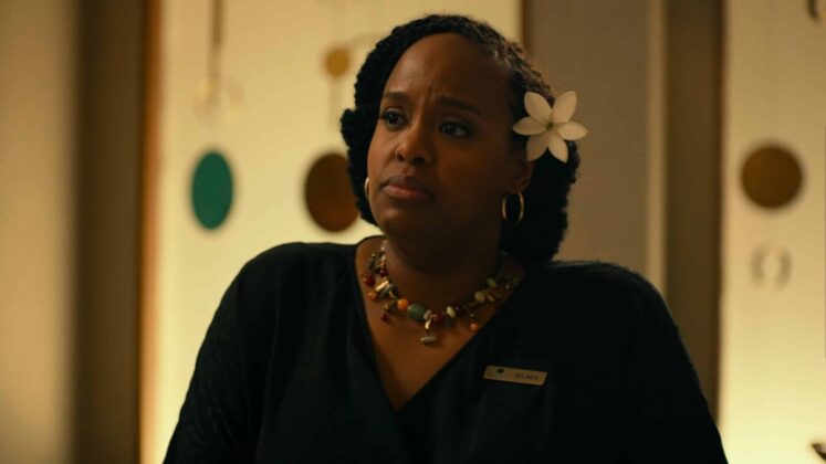 Na imagem vê-se uma mulher preta com uniforme preto, colar volumoso e flor havaiana na cabeça.