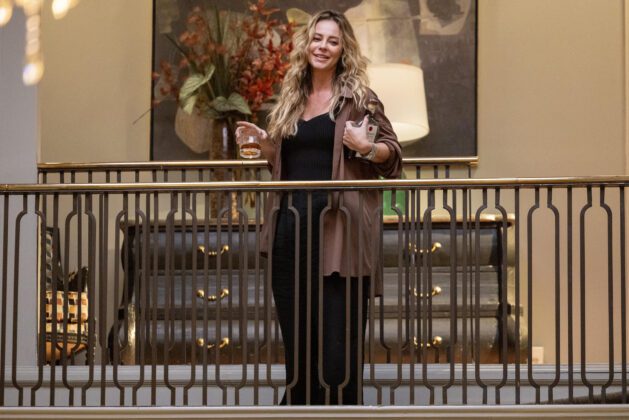 Paolla Oliveira como Heleninha no alto da escada. Ela segura um copo de bebida alcoólica