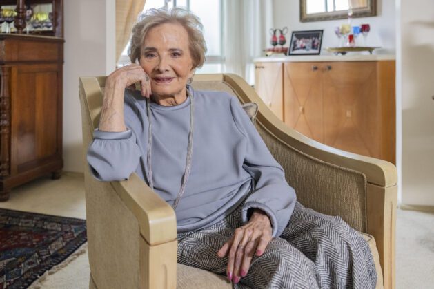 Glória Menezes sentada numa poltrona na sala de sua casa